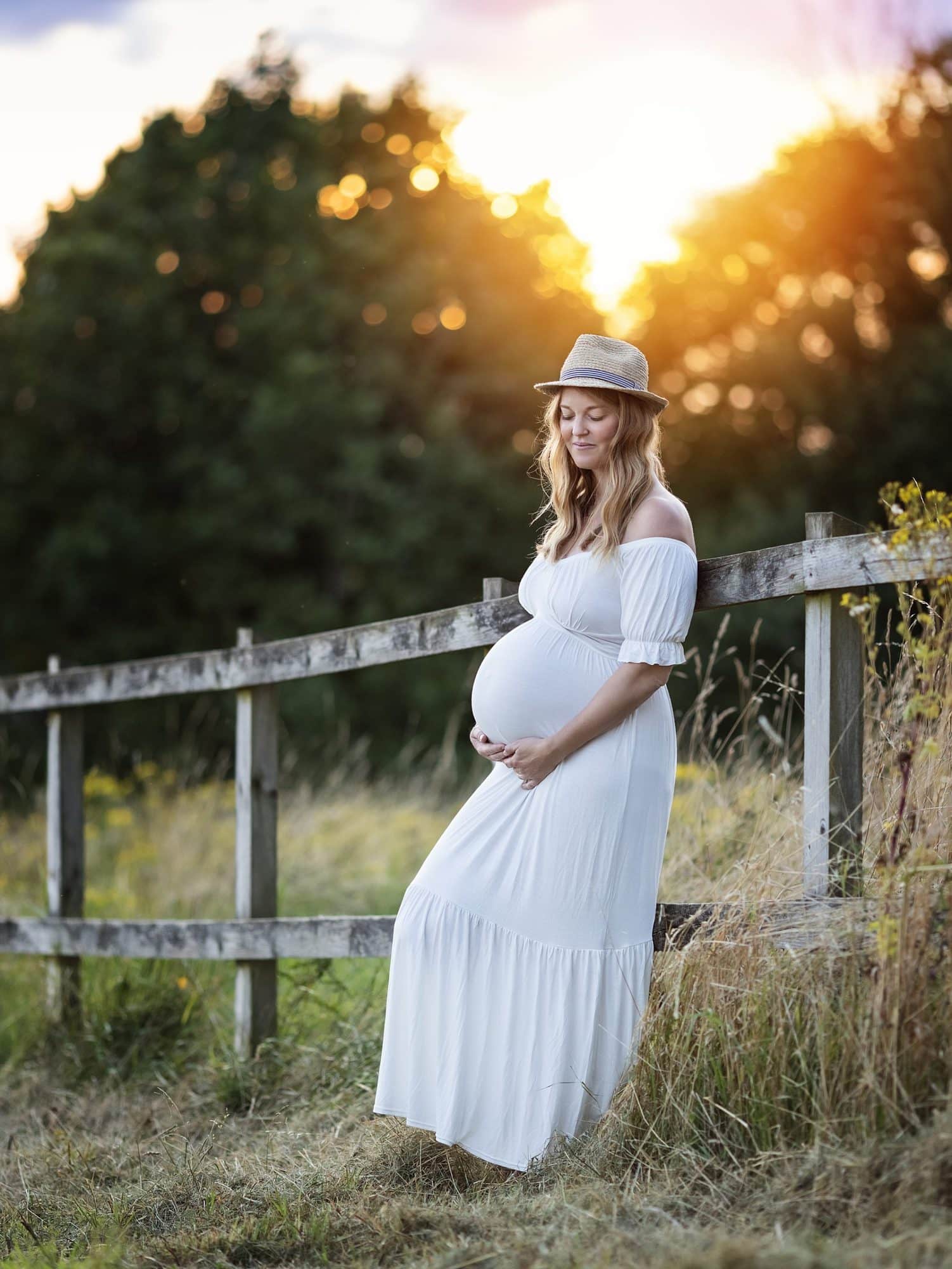 Pregnant woman leans on fence with sunset in background