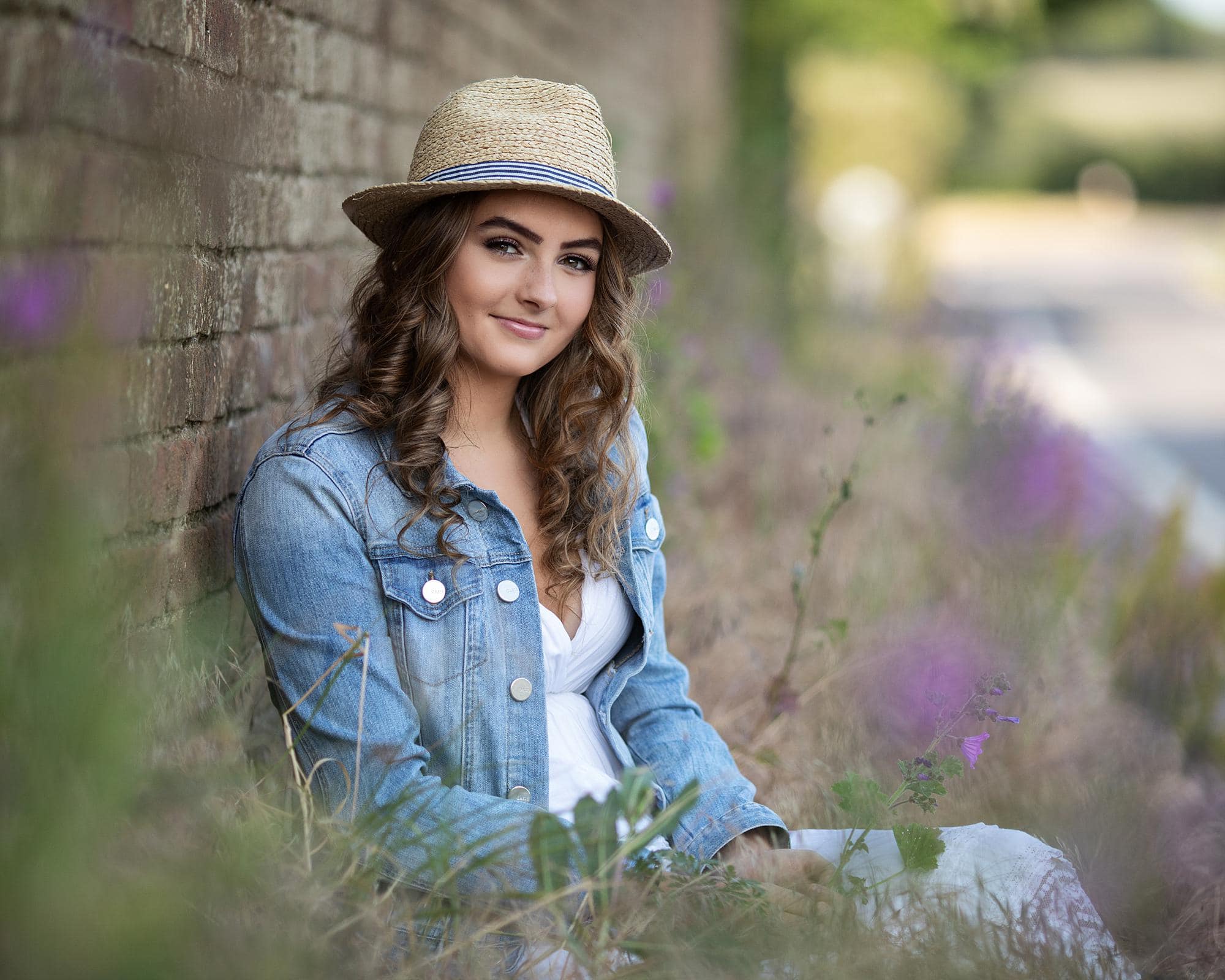 Smiling teenage girl sits wearing a straw hat, white dress and denim jacket for an outdoor family photoshoot in Suffolk