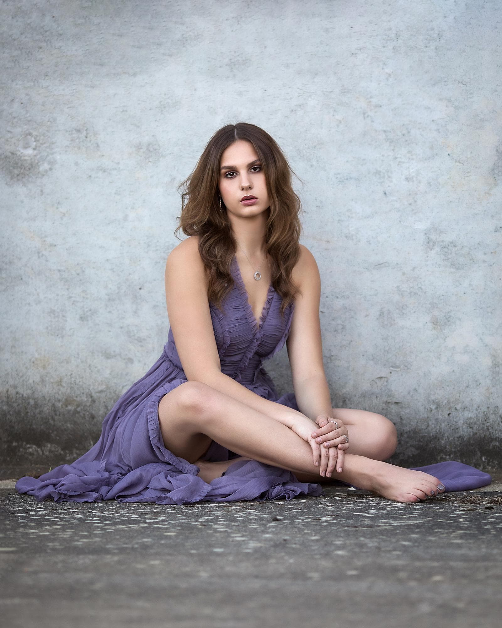 Woman in a purply dress sits on the ground and against a concrete wall