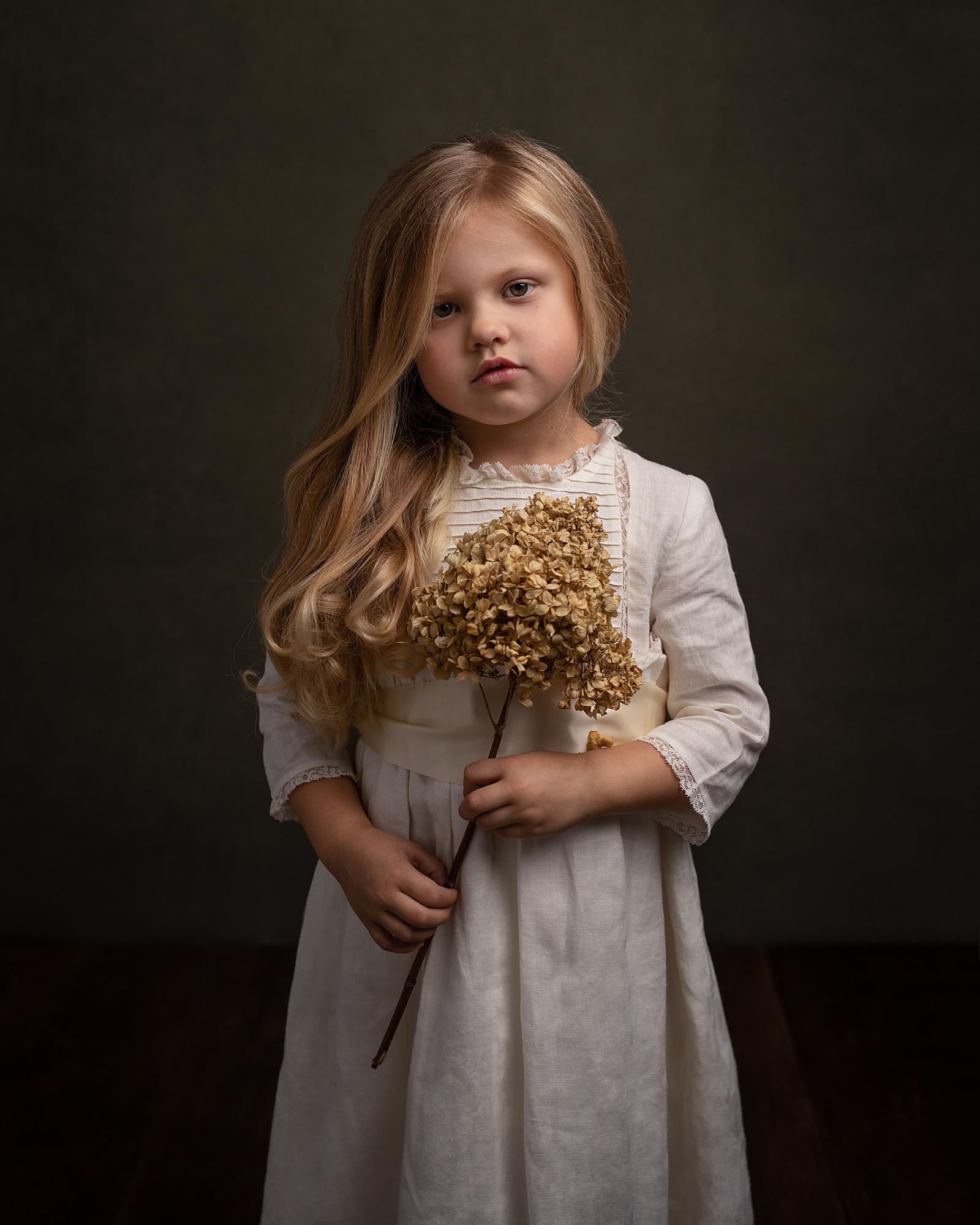 Little girl holds a dried flower wearing a vintage cream dress in a Fine Art Photoshoot in Suffolk