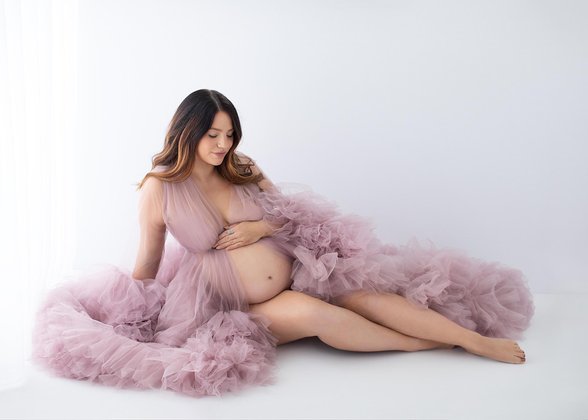 Pregnant woman posing against a white backdrop with a sheer pink gown for a maternity photoshoot in Suffolk photography studio