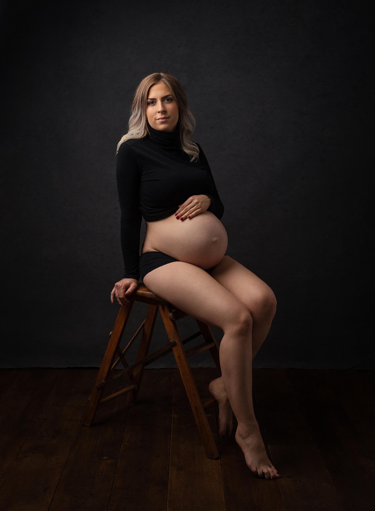Pregnant woman sits on a stool posing against a black backdrop for a maternity photoshoot in Suffolk photography studio