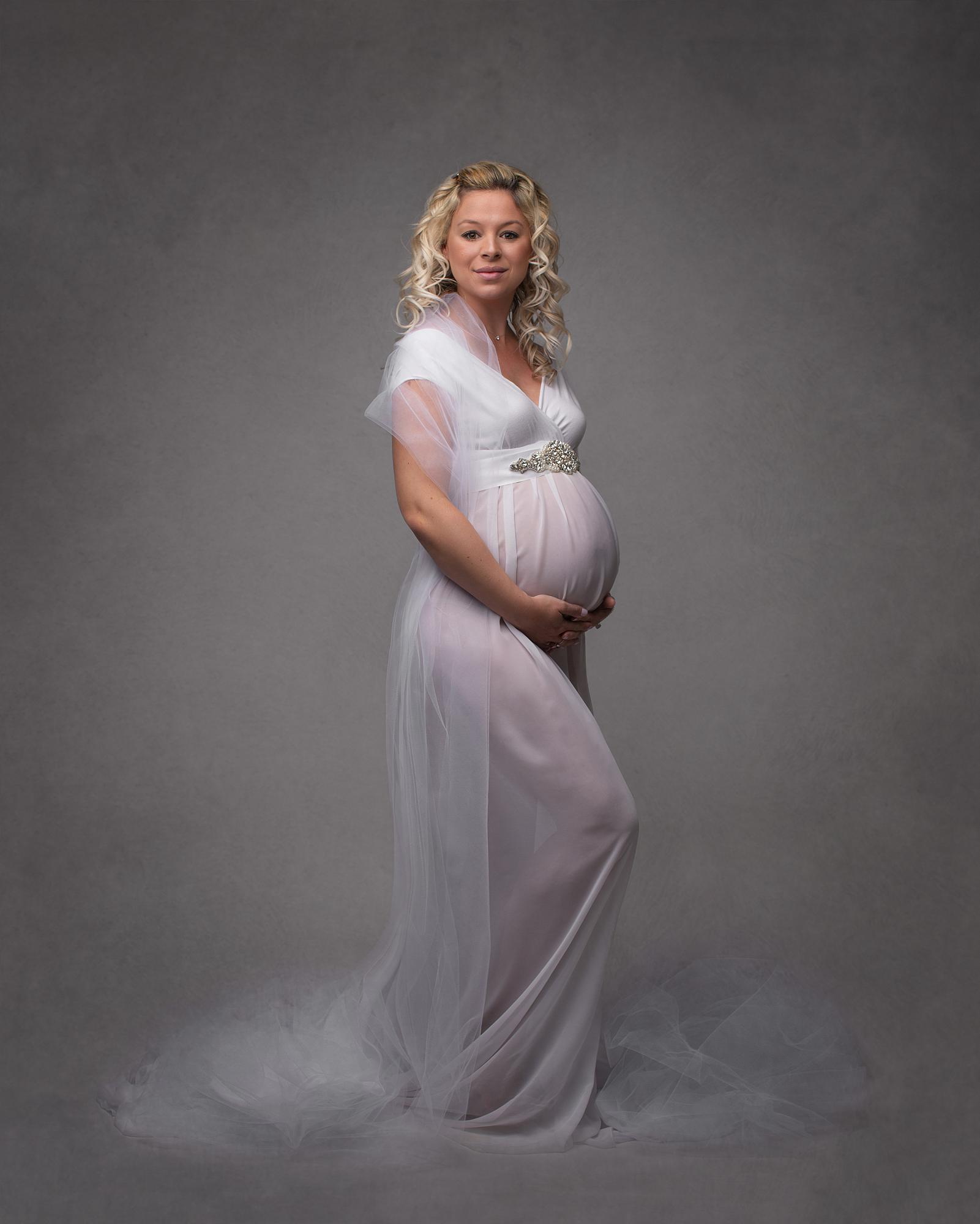 Pregnant woman posing in a white dress against a grey backdrop for a maternity photoshoot in Suffolk photography studio
