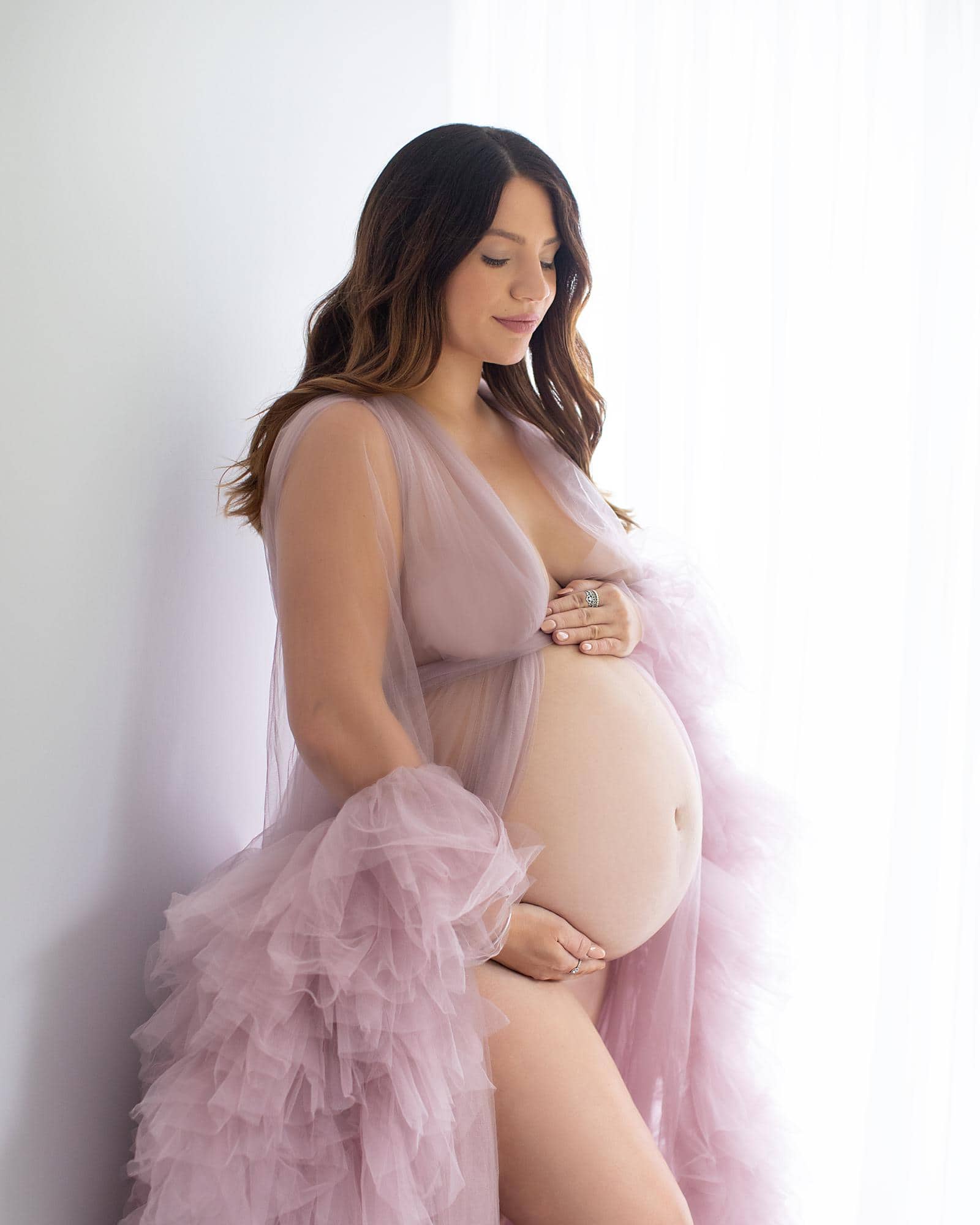 Pregnant woman posing against a white backdrop with a sheer pink gown for a maternity photoshoot in Suffolk photography studio