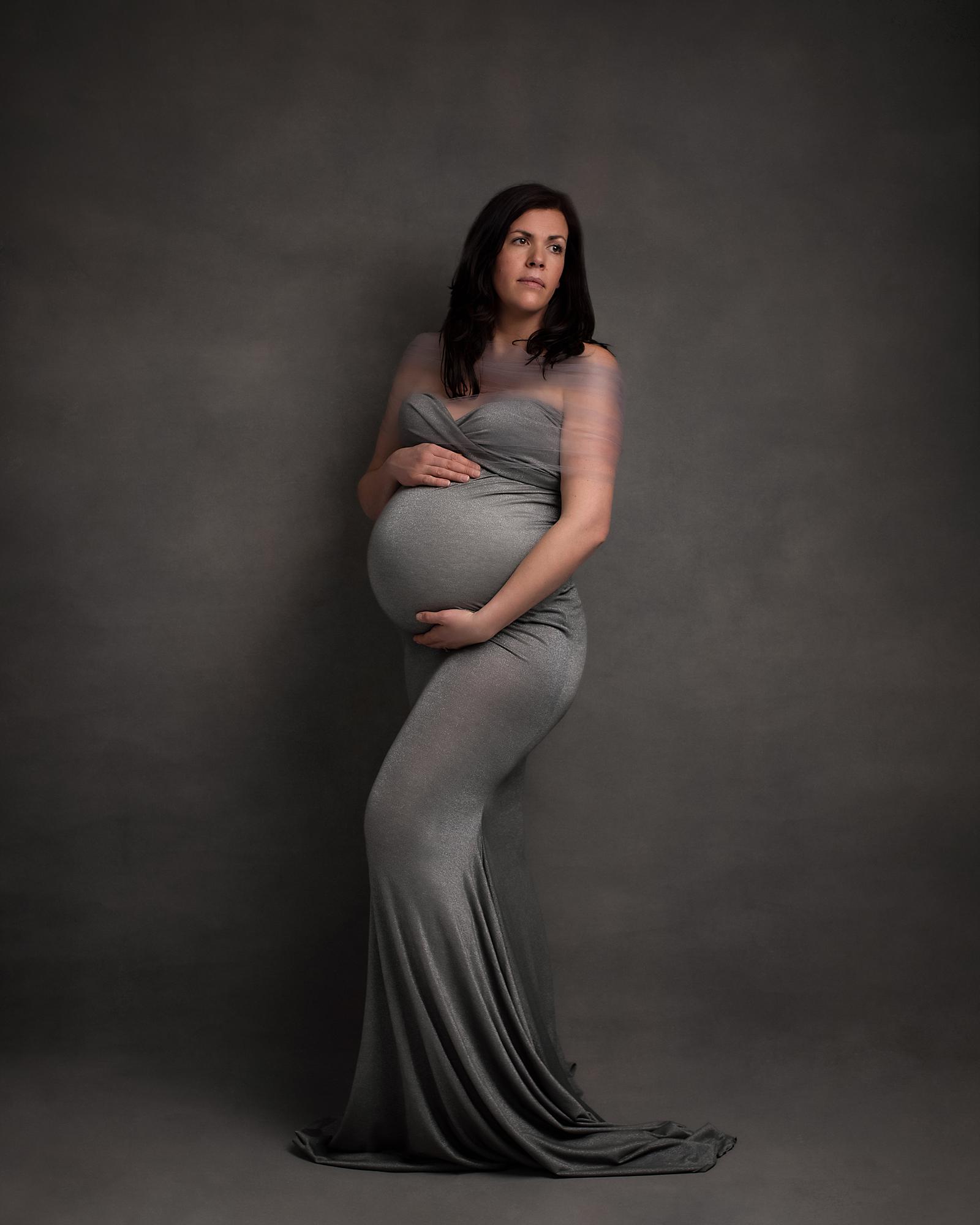 Pregnant woman posing in a grey dress against a grey backdrop for a maternity photoshoot in Suffolk