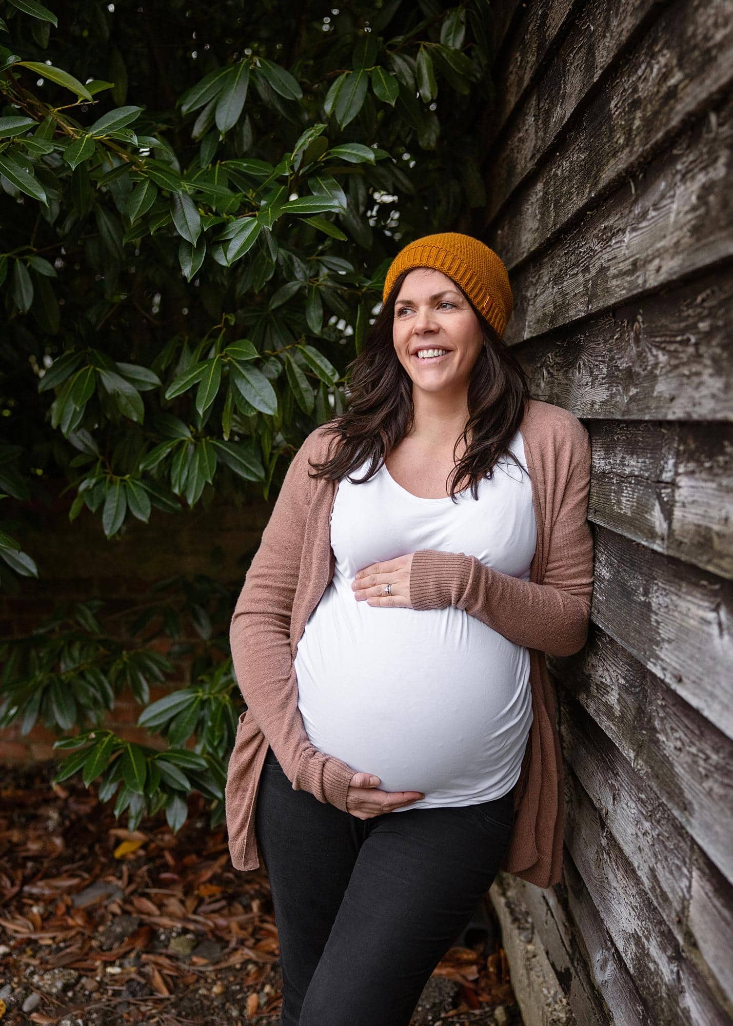 Pregnant woman posing in a white vest and yellow hat leaning against a barn door for a maternity photography shoot in Suffolk
