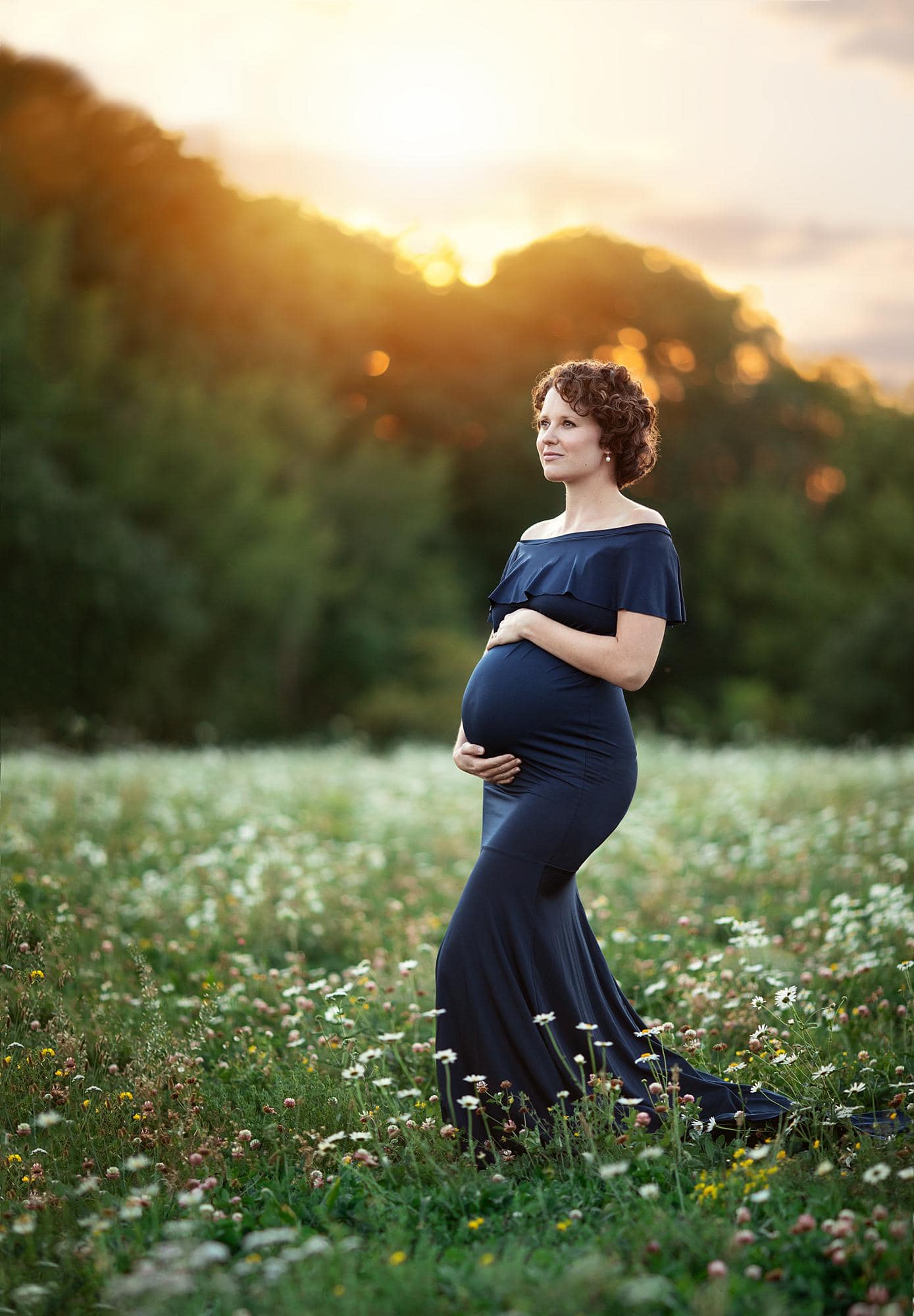 Woman smiles while holding her pregnant belly and posing for a maternity photoshoot in Suffolk flower field while the sun sets in the background