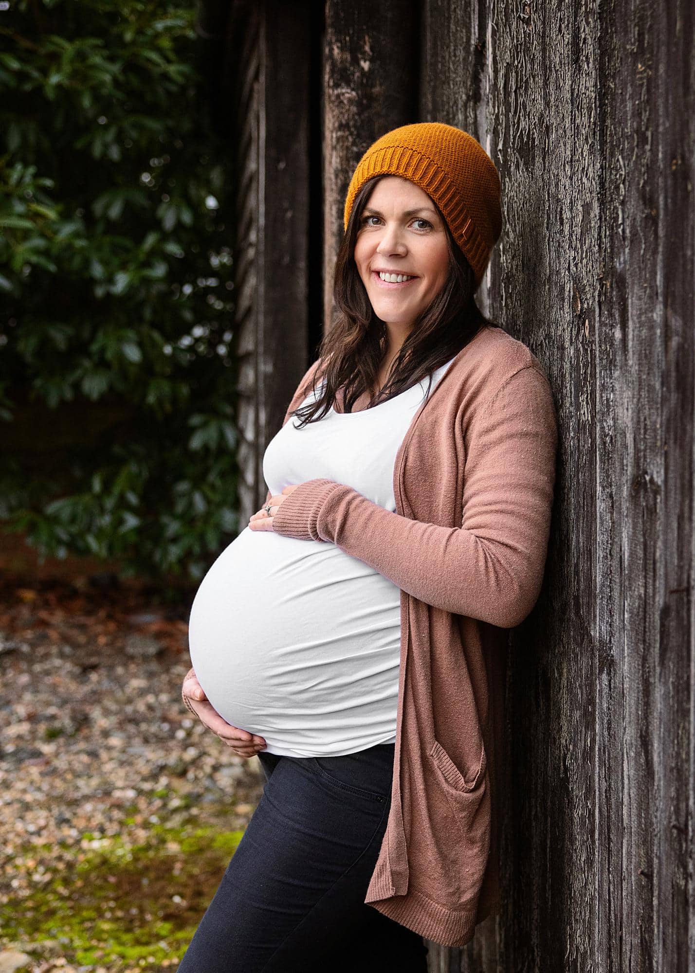 Pregnant woman posing in a white vest and yellow hat leaning against a barn door for a maternity photography shoot in Suffolk