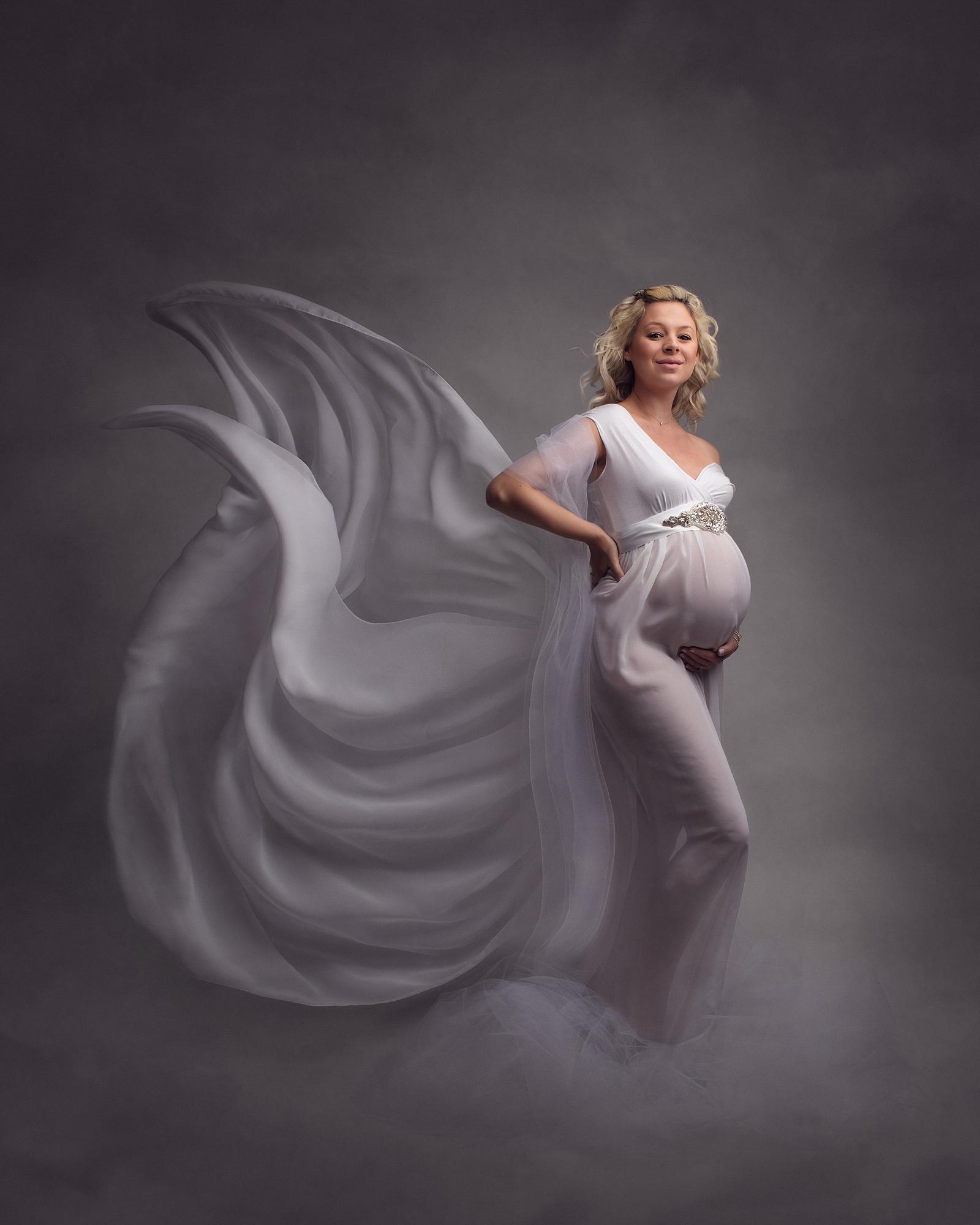 Pregnant woman posing in sheer white dress with flowing material for a maternity photoshoot in Alison McKenny's Suffolk studio