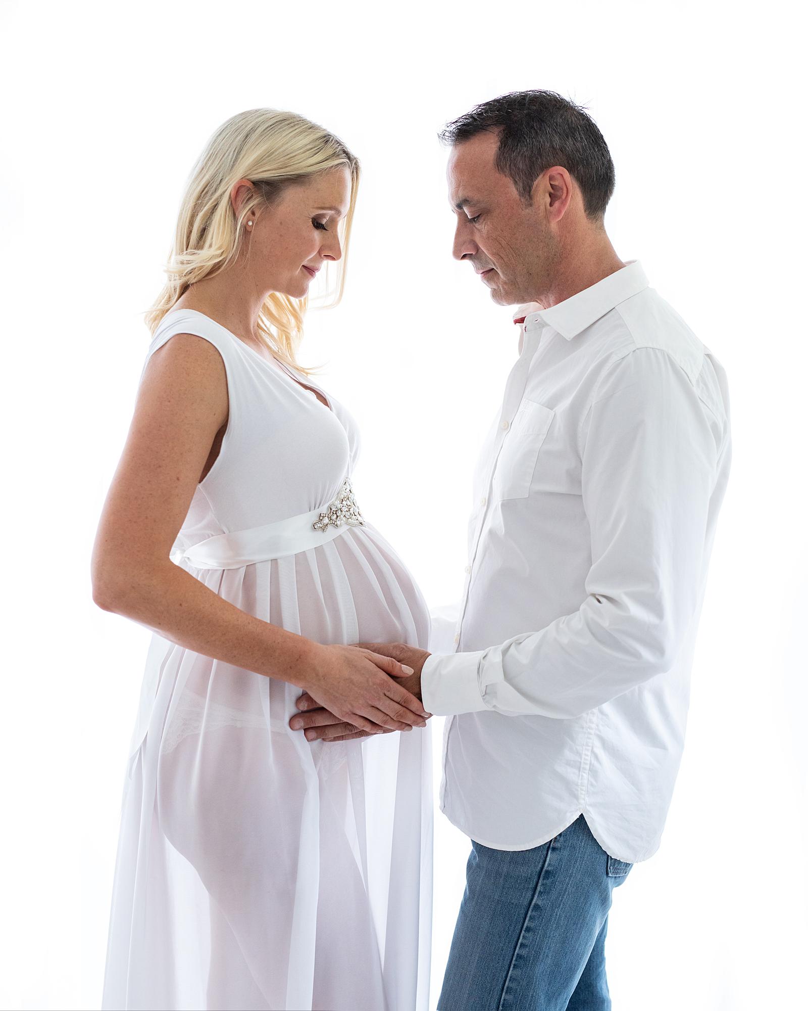 Pregnant woman posing in sheer white dress with her partner touching her belly for a maternity photoshoot in Suffolk
