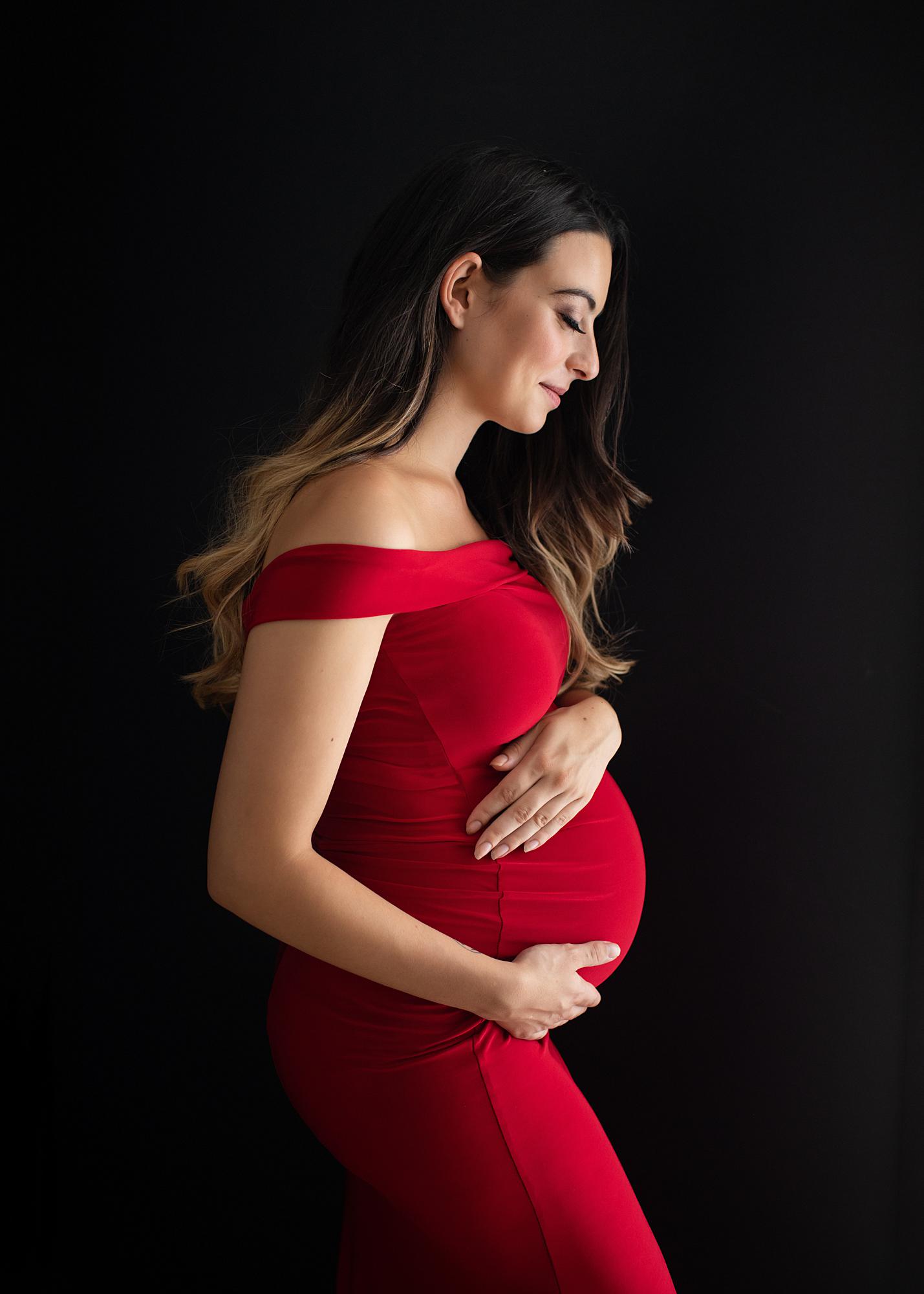 Pregnant woman posing in a red dress holds her pregnant belly for a maternity photoshoot in Suffolk