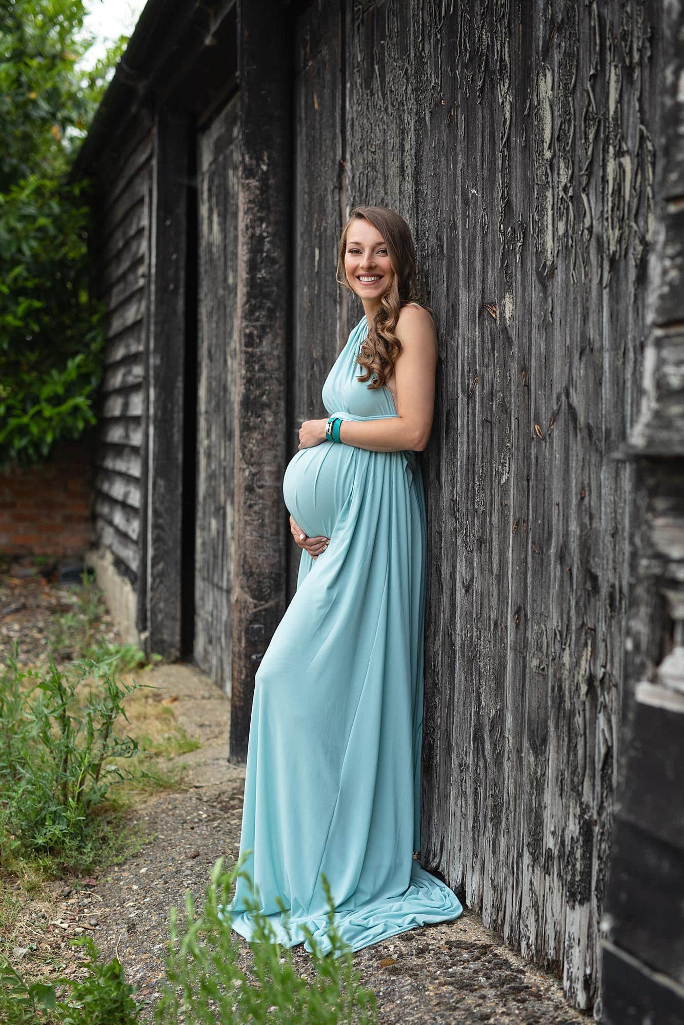 Pregnant woman posing in a blue dress against a barn door for a maternity photoshoot in Alison McKenny's Suffolk studio
