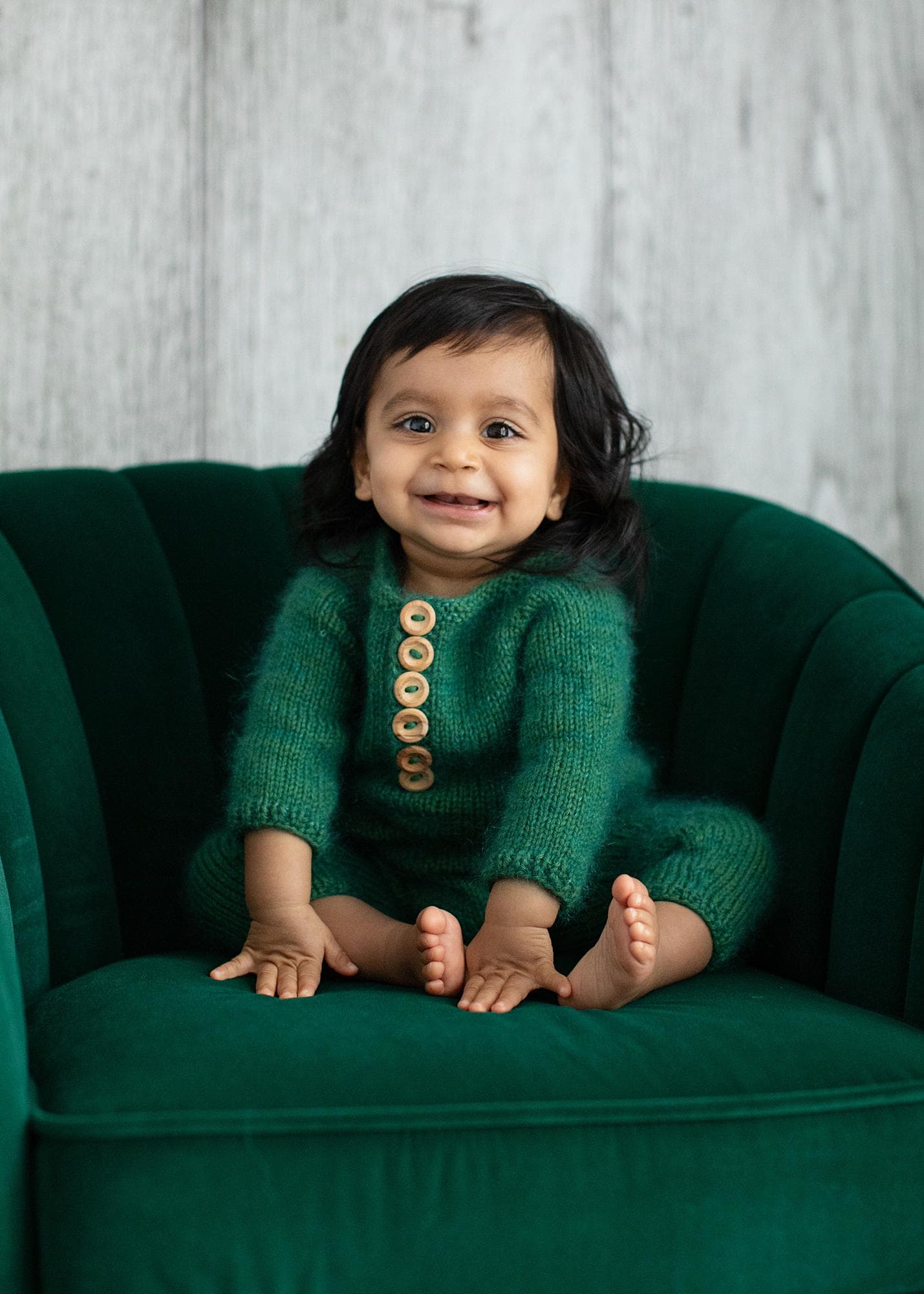 Baby Girl with long black hair sits smiling on a green velvet chair for Baby Photo Shoot