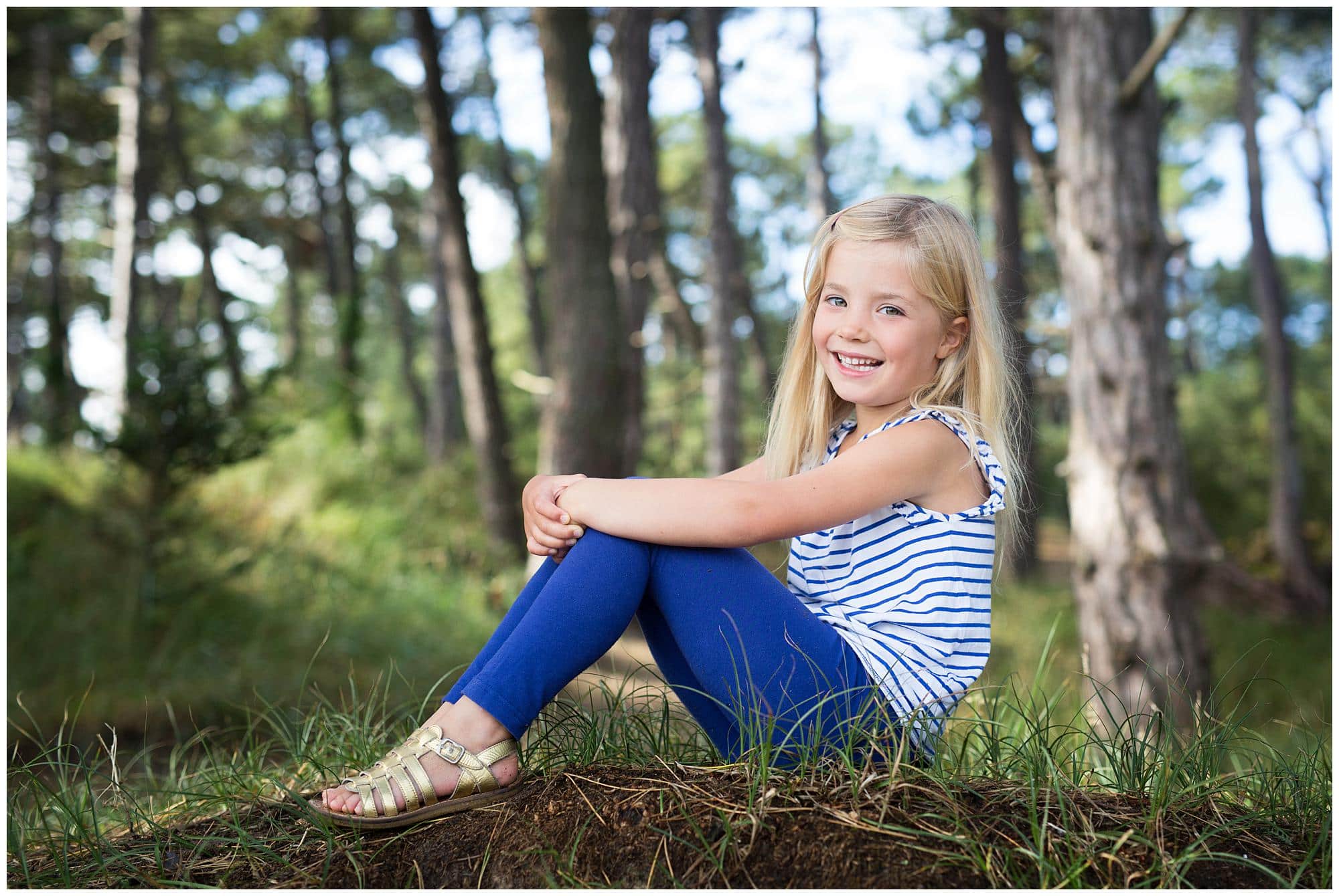 Little girl in blue and white striped top sitting in woods in Suffolk and smiling