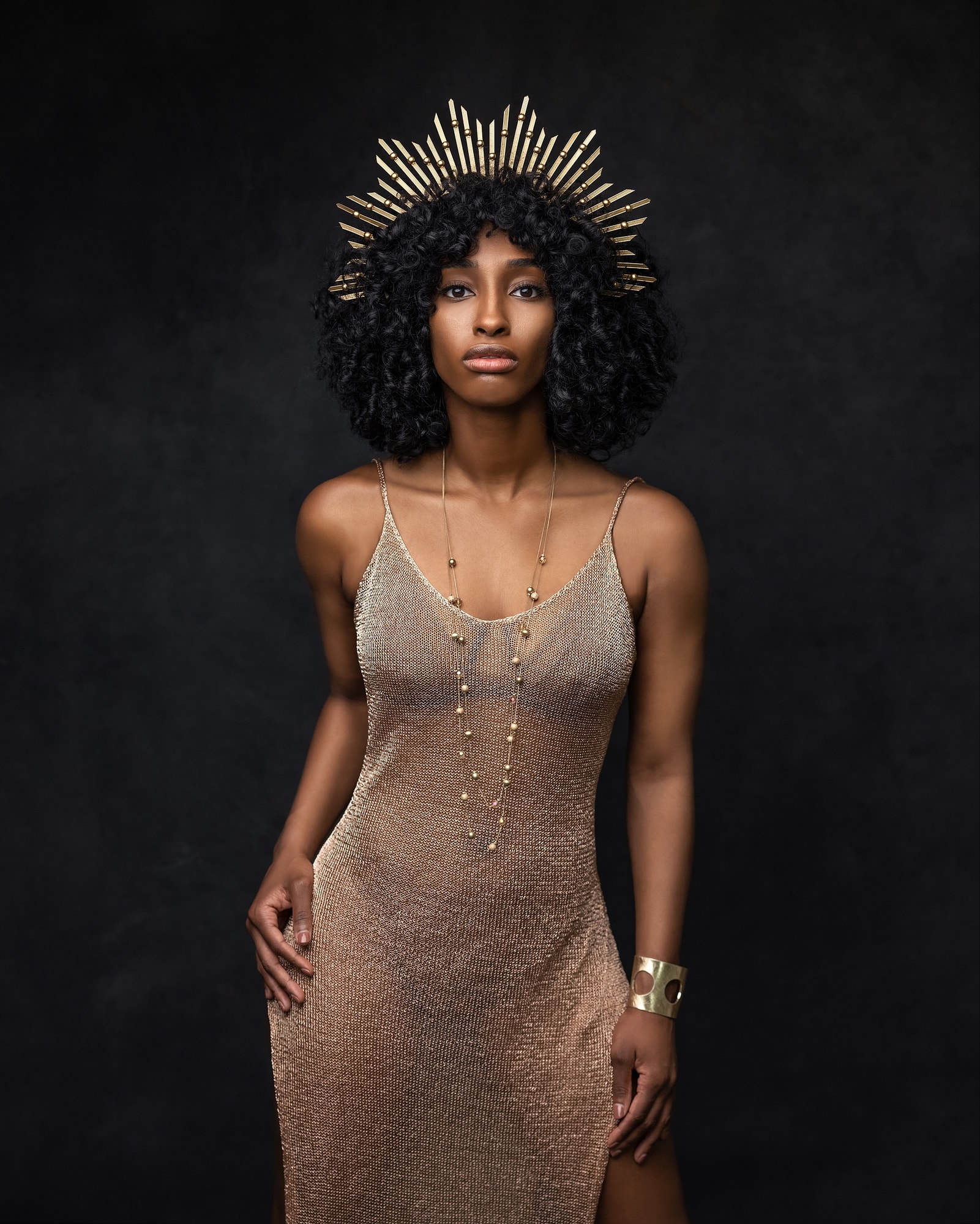 Beautiful woman in a rose gold dress and gold crown poses for a beauty shoot on a black background in Suffolk studio