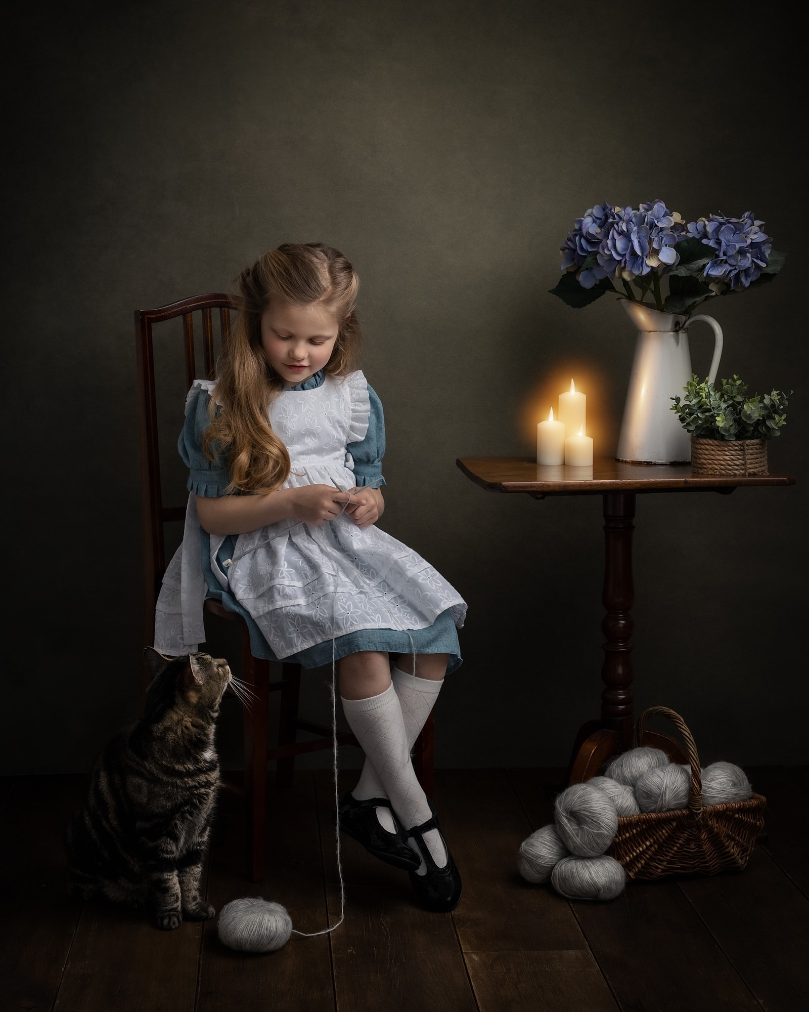 Little girl wearing a blue dress with white apron sits knitting against a dark background with her cat looking at her during fine art photoshoot at Suffolk Studio