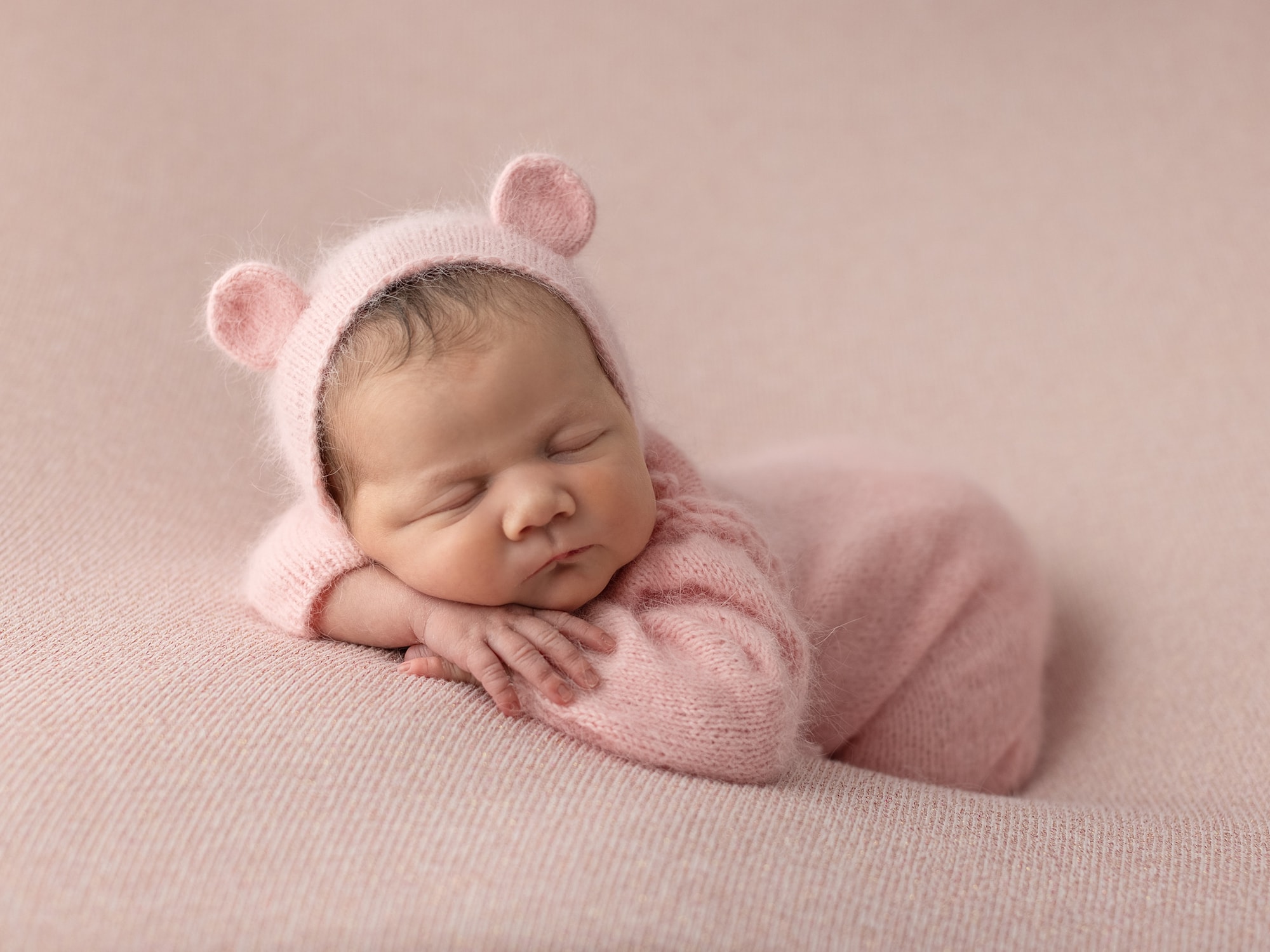 Newborn Baby Girl wearing a pink wool romper and pink teddy bear bonnet on a pink blanket