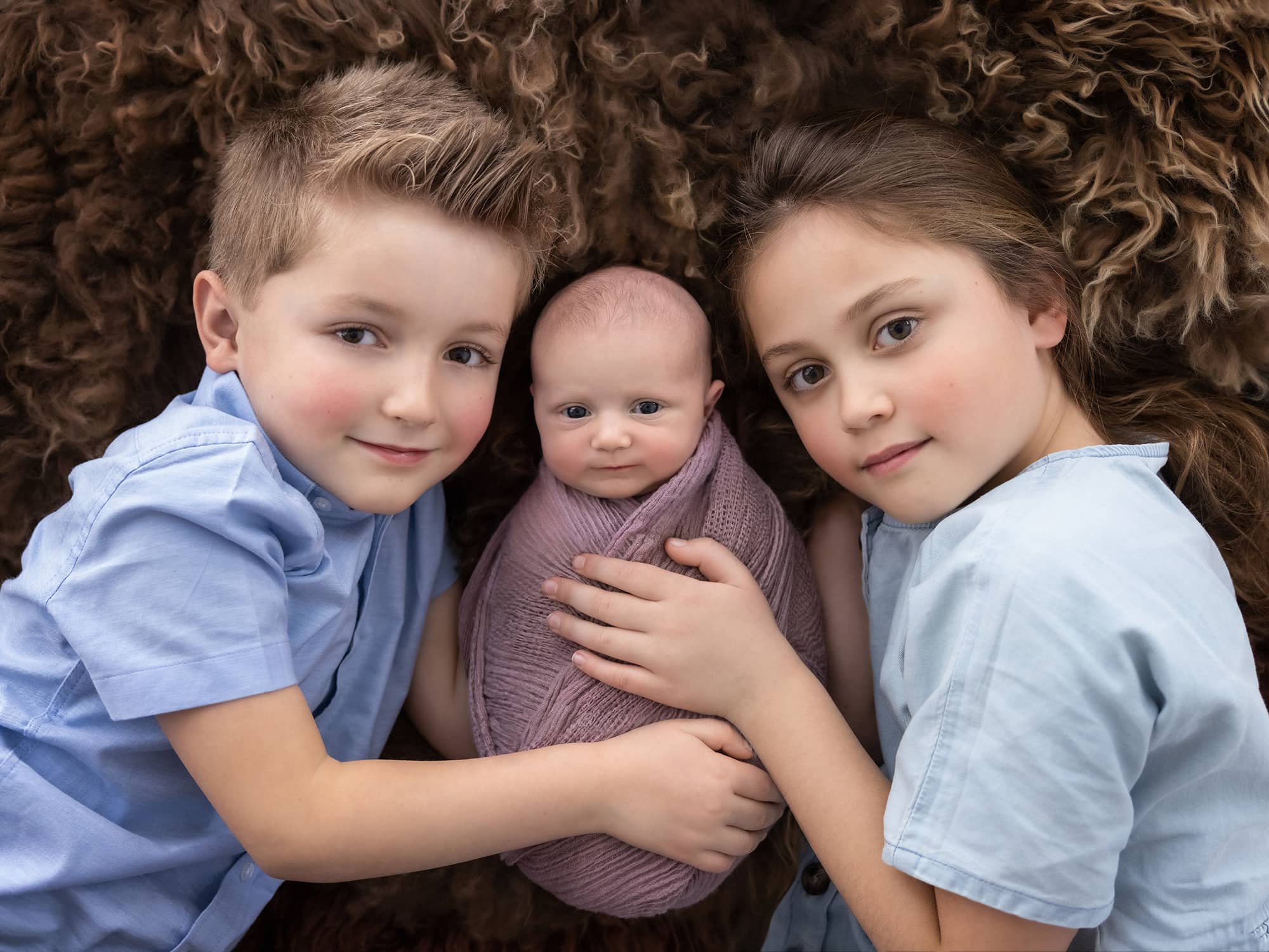 A brother and Sister cuddle their newborn baby sister on a brown rug