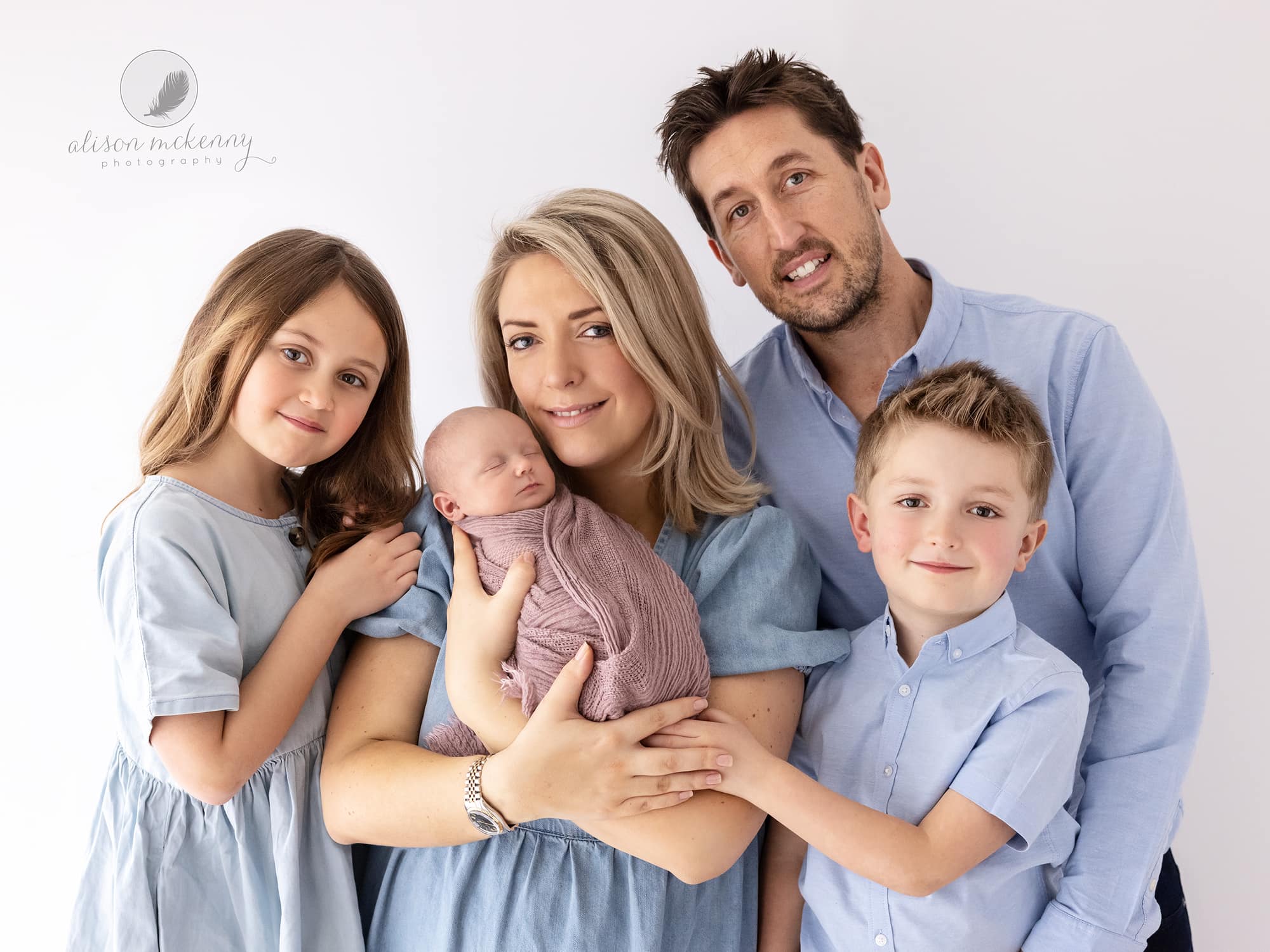 Family of 5 on a white background wear blue and Mother holds newborn baby girl in a pink blanket