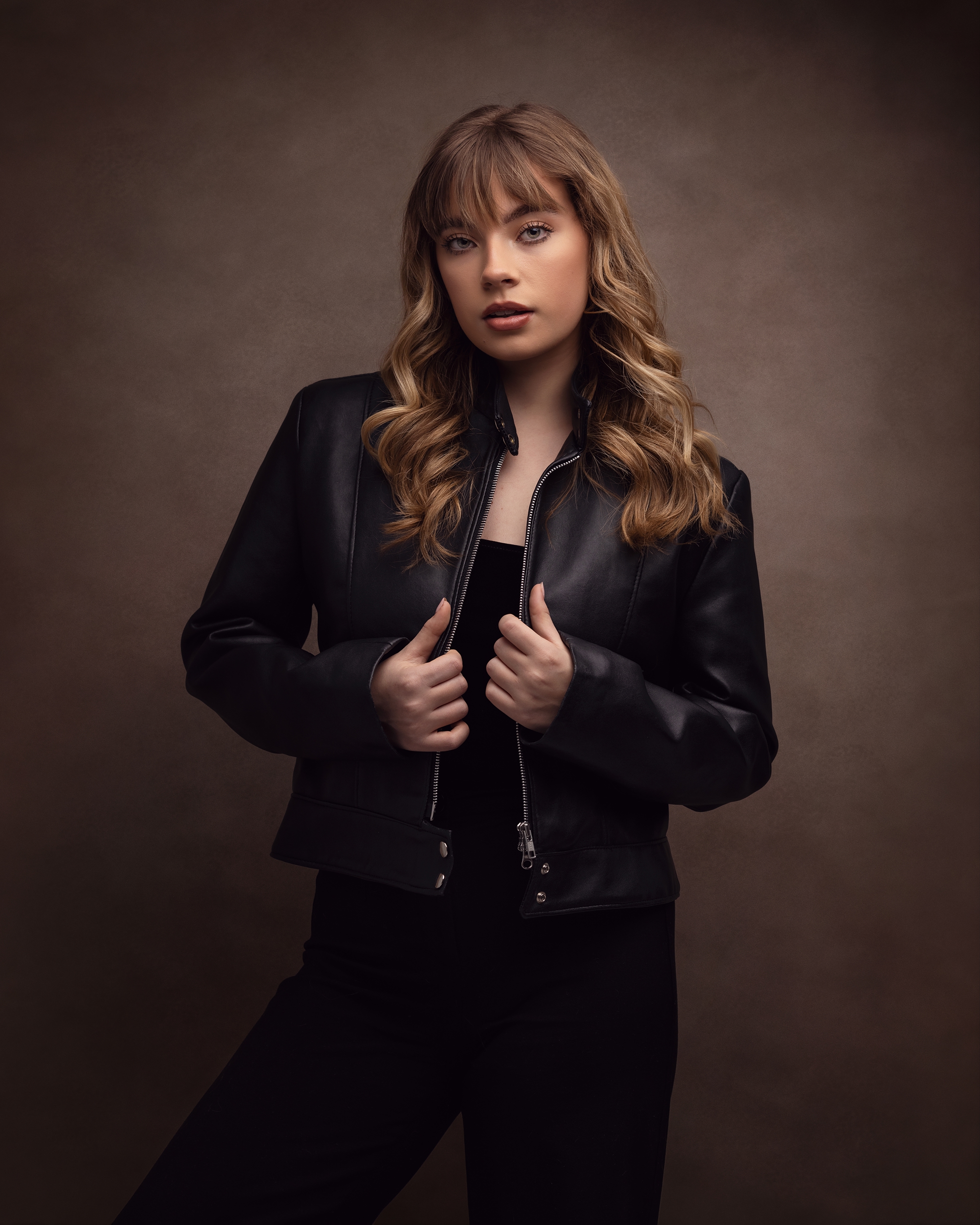 Teenage girl in a black leather jacket and trousers poses against a brown background