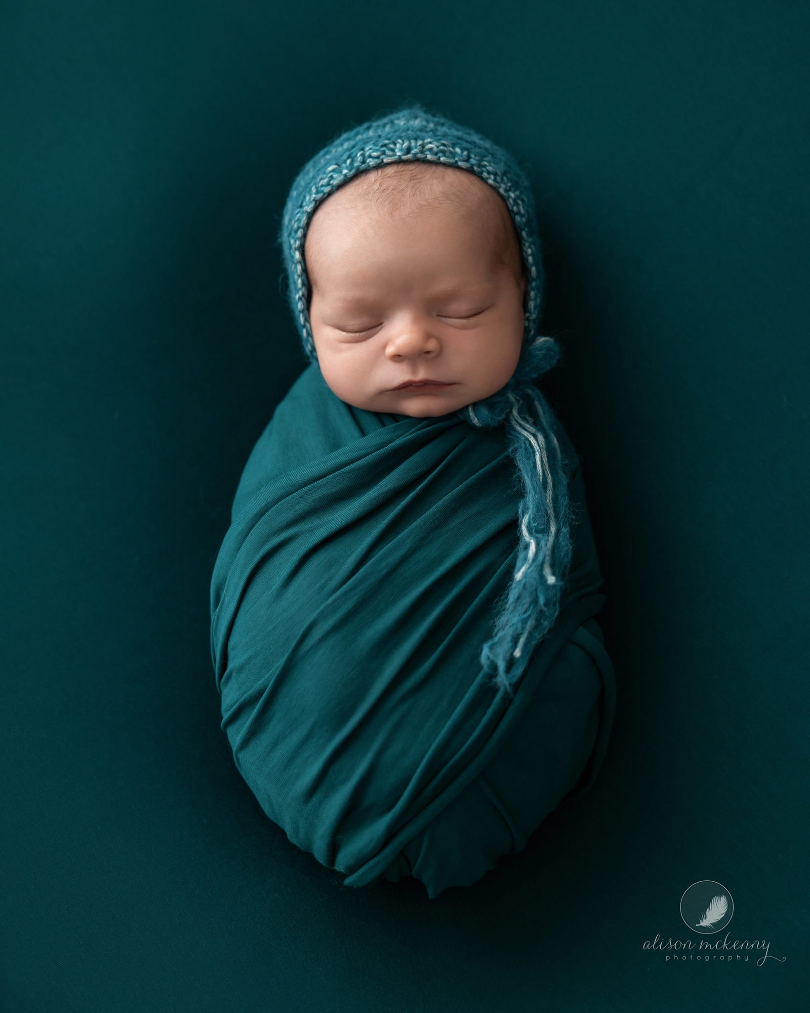 Newborn Baby Boy wrapped in a teal wrap, lies on a teal blanket and has a teal bonnet on his head. He sleeps peacefully during his newborn shoot with suffolk photographer Alison McKenny