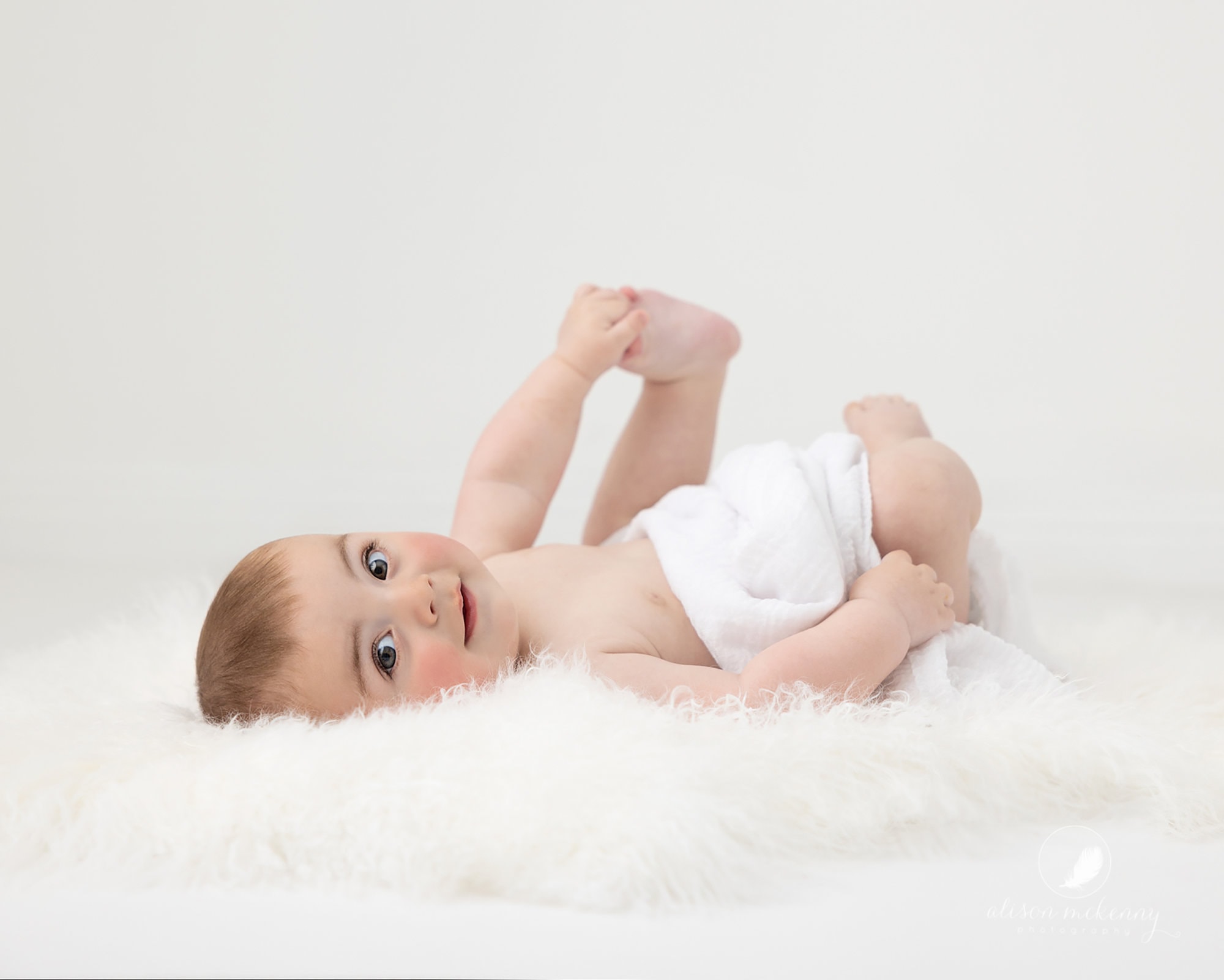 6 month old baby boy holds his feet and smiles at camera during Baby Photoshoot in Suffolk Studio