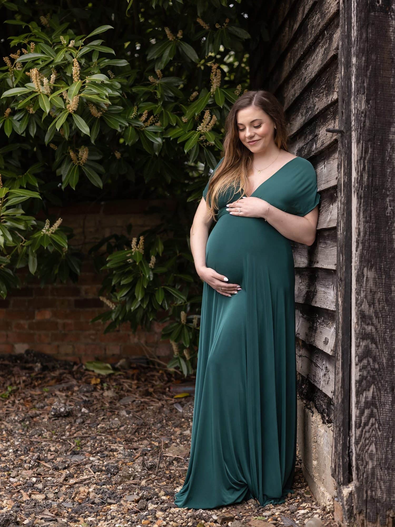 Pregnant lady in green dress leans against old barn during Maternity shoot on Suffolk farm