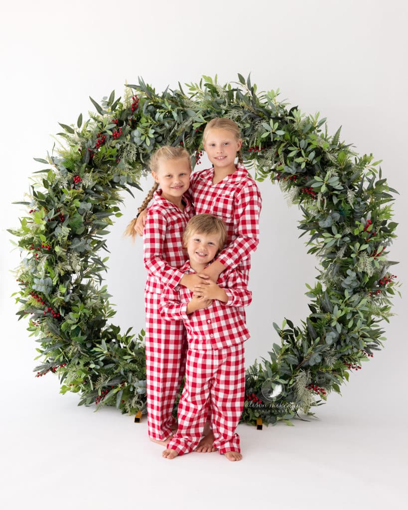 3 Siblings wearing red and white pyjamas hug each other during Christmas Mini Session in Suffolk Studio