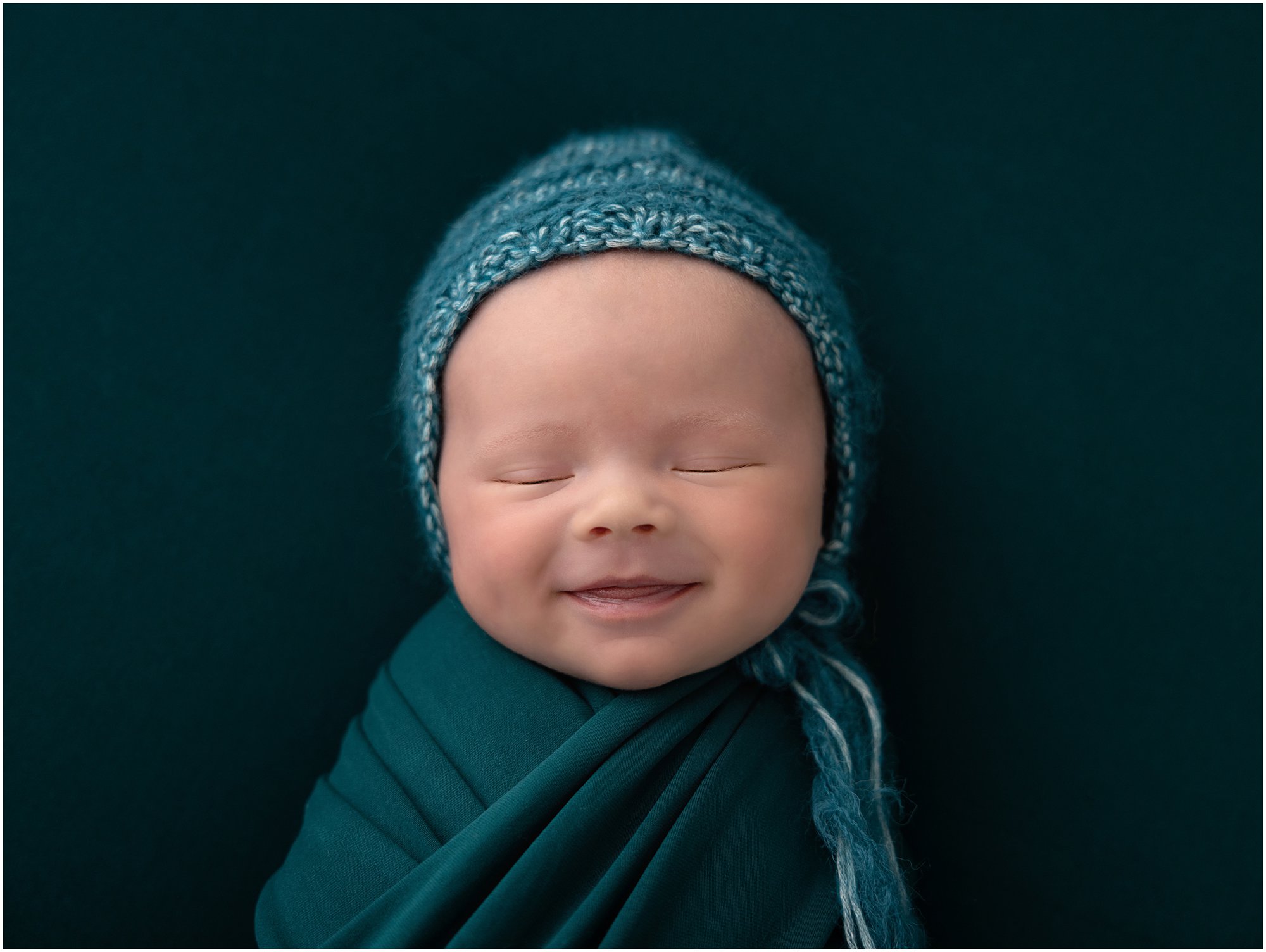 Newborn baby smiles while wearing an emerald green bonnet and wrap on a emerald green blanket