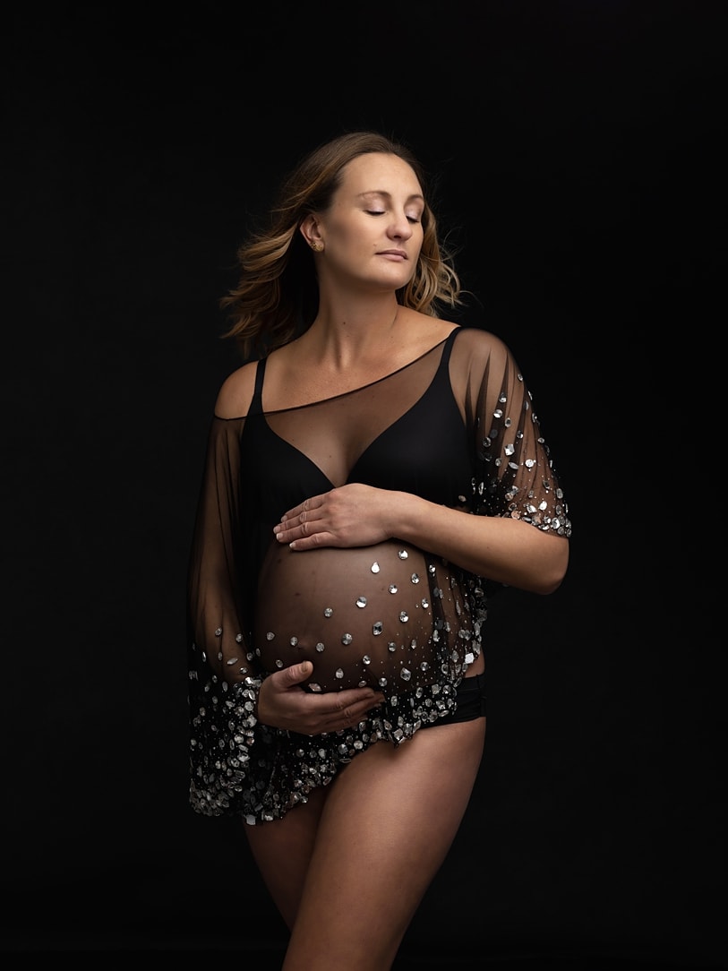 Beautiful pregnant lady closes her eyes and holds her bump while posing in her black underwear and a black sheer cape during a Maternity Photoshoot with Alison McKenny in her studio near Clare, Suffolk
