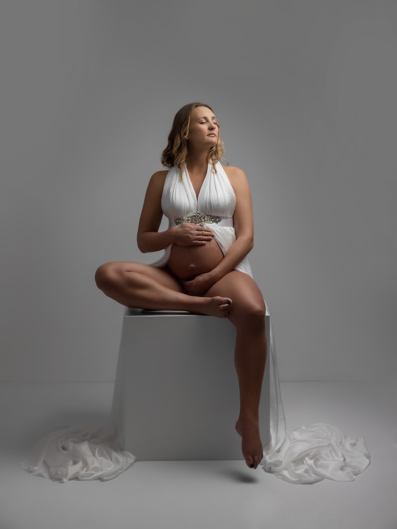Beautiful pregnant lady closes her eyes and holds her bump while wearing a white dress and sitting on a white box during a Maternity Photoshoot with Alison McKenny in her studio near Clare, Suffolk