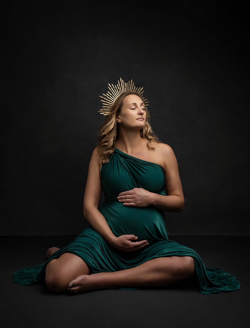 Beautiful pregnant lady closes her eyes and holds her bump while wearing a green dress and gold crown posing during a Maternity Photoshoot with Alison McKenny in her studio near Clare, Suffolk