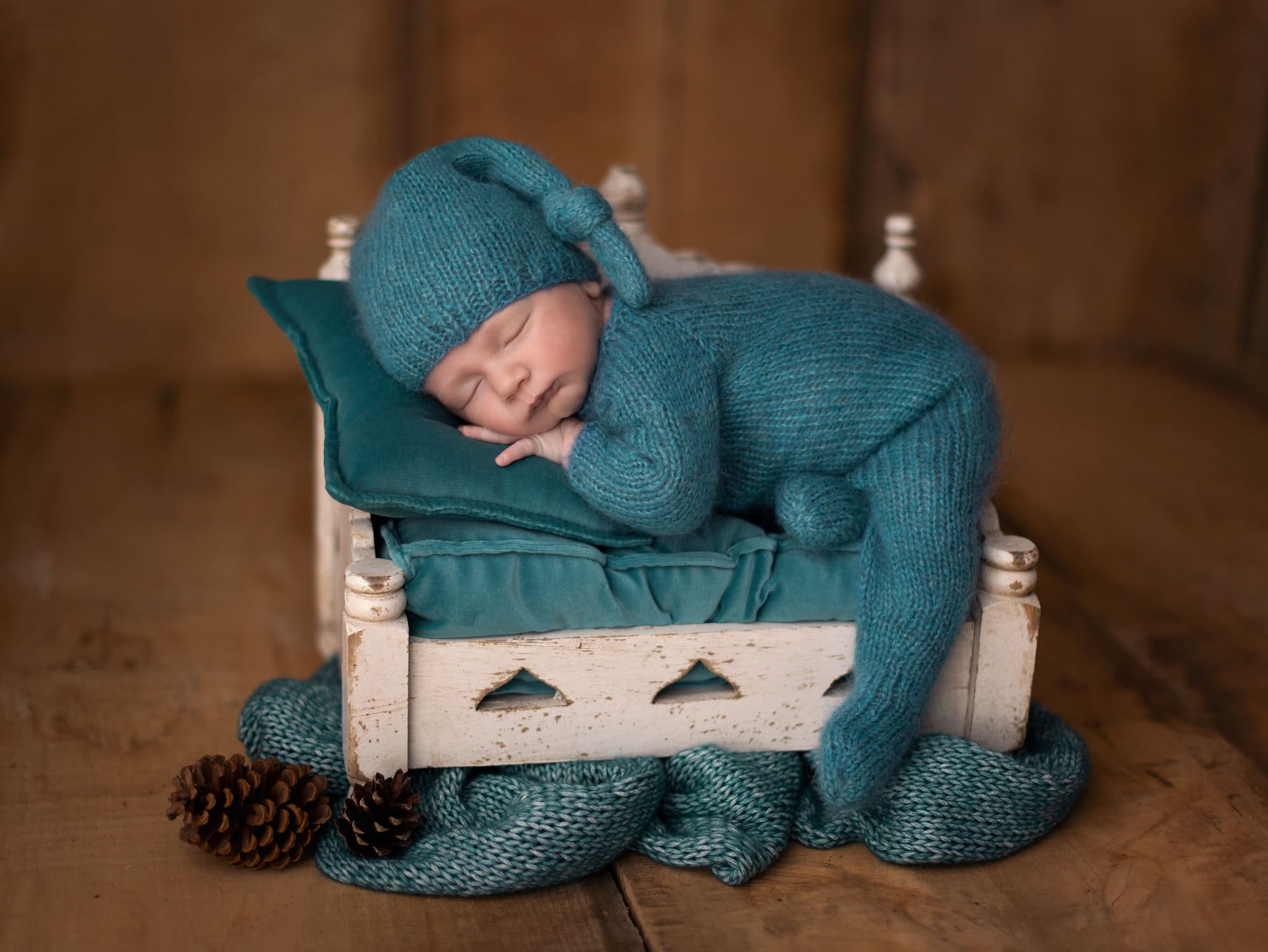 Newborn baby boy in a teal wool romper and hat sleeps on a Newborn bed on a brown wooden floor during his first photoshoot with Alison McKenny, Cambridge Photographer