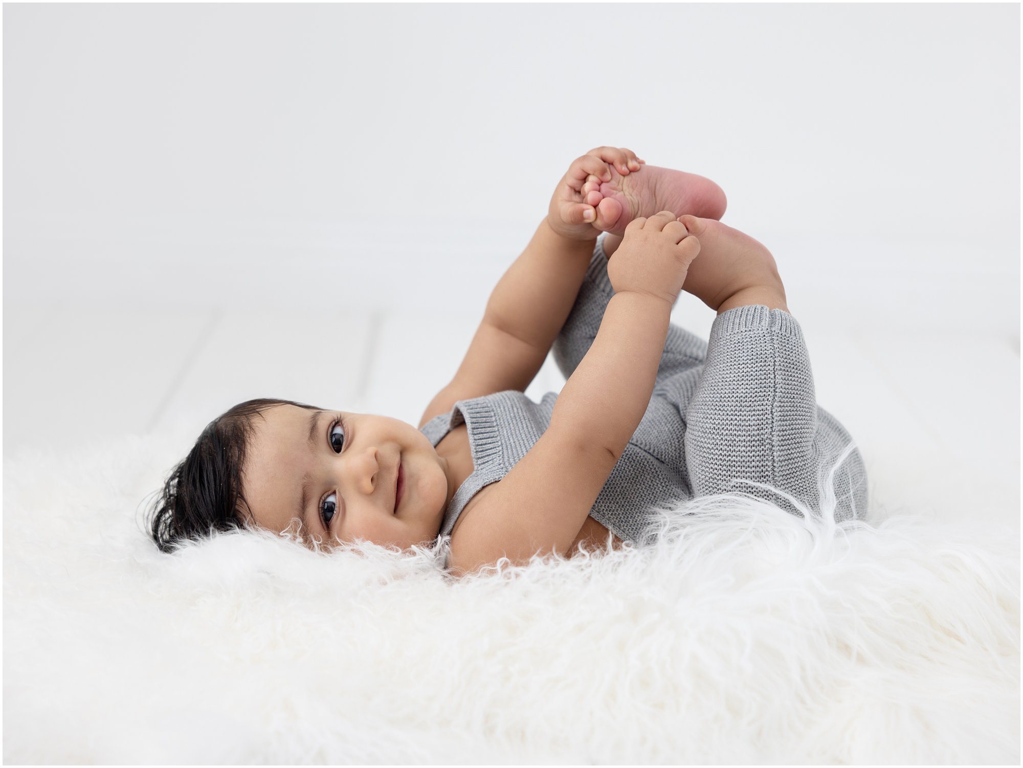 Cute baby grabs his toes as he lays on a white rug and smiles at camera during his Baby Photoshoot