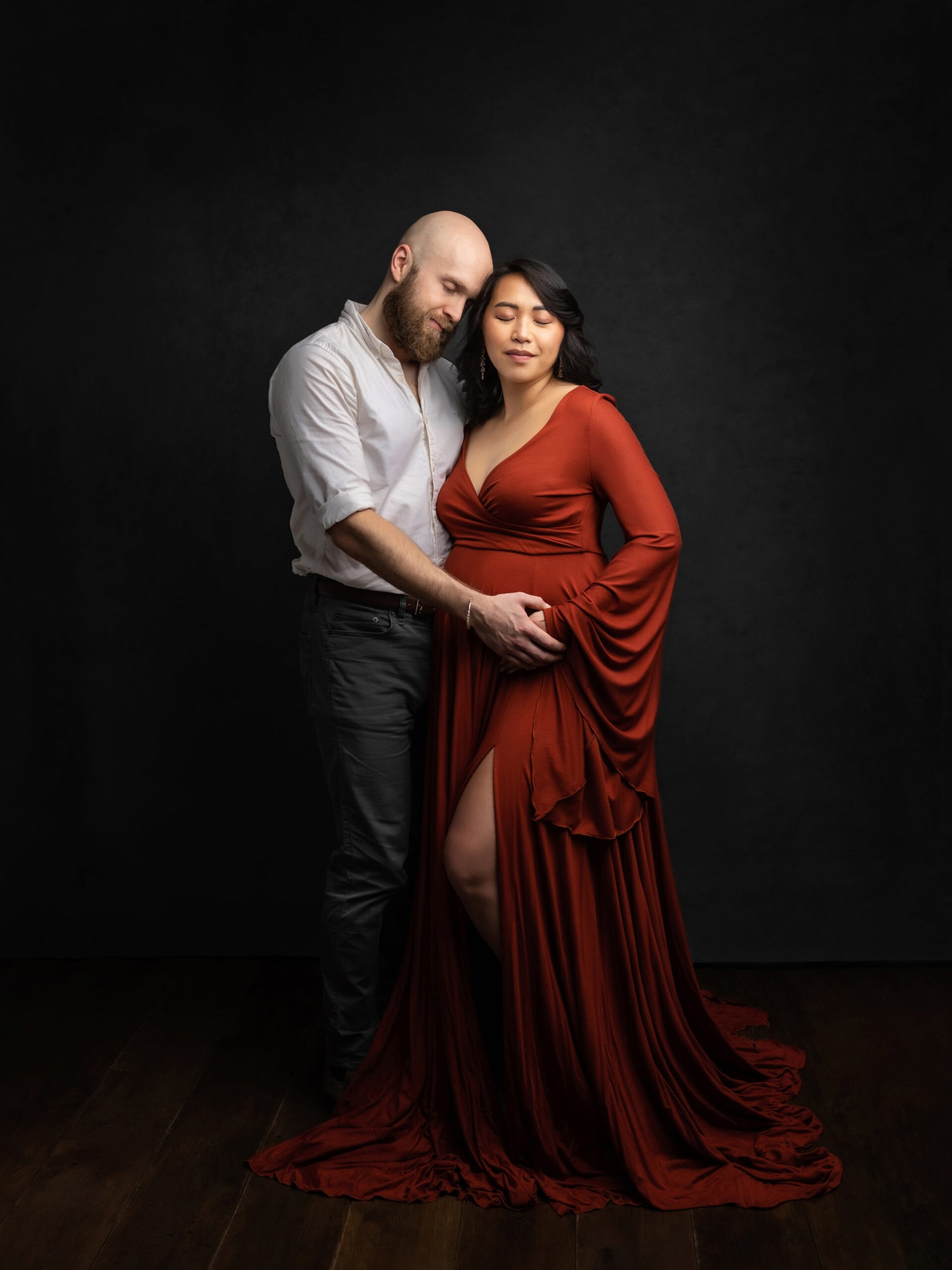 Pregnant lady in a rust coloured dress embraces her husband as they touch her bump during Maternity Shoot in Suffolk studio