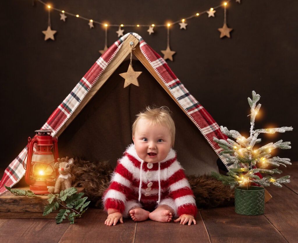 Baby boy in red and white wool romper sits under a tent on a Christmas Mini Shoot set in suffolk studio