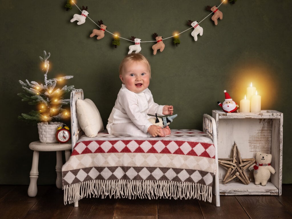 Baby boy in red and white wool romper sits on a small bed on a Christmas Mini Shoot set in suffolk studio