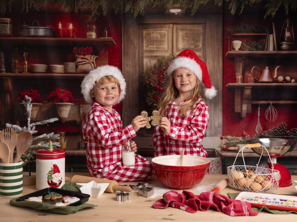 Two kids in red and white pj's smile at the camera during their Christmas Mini Session in a festive kitchen set up with Alison McKenny Photography