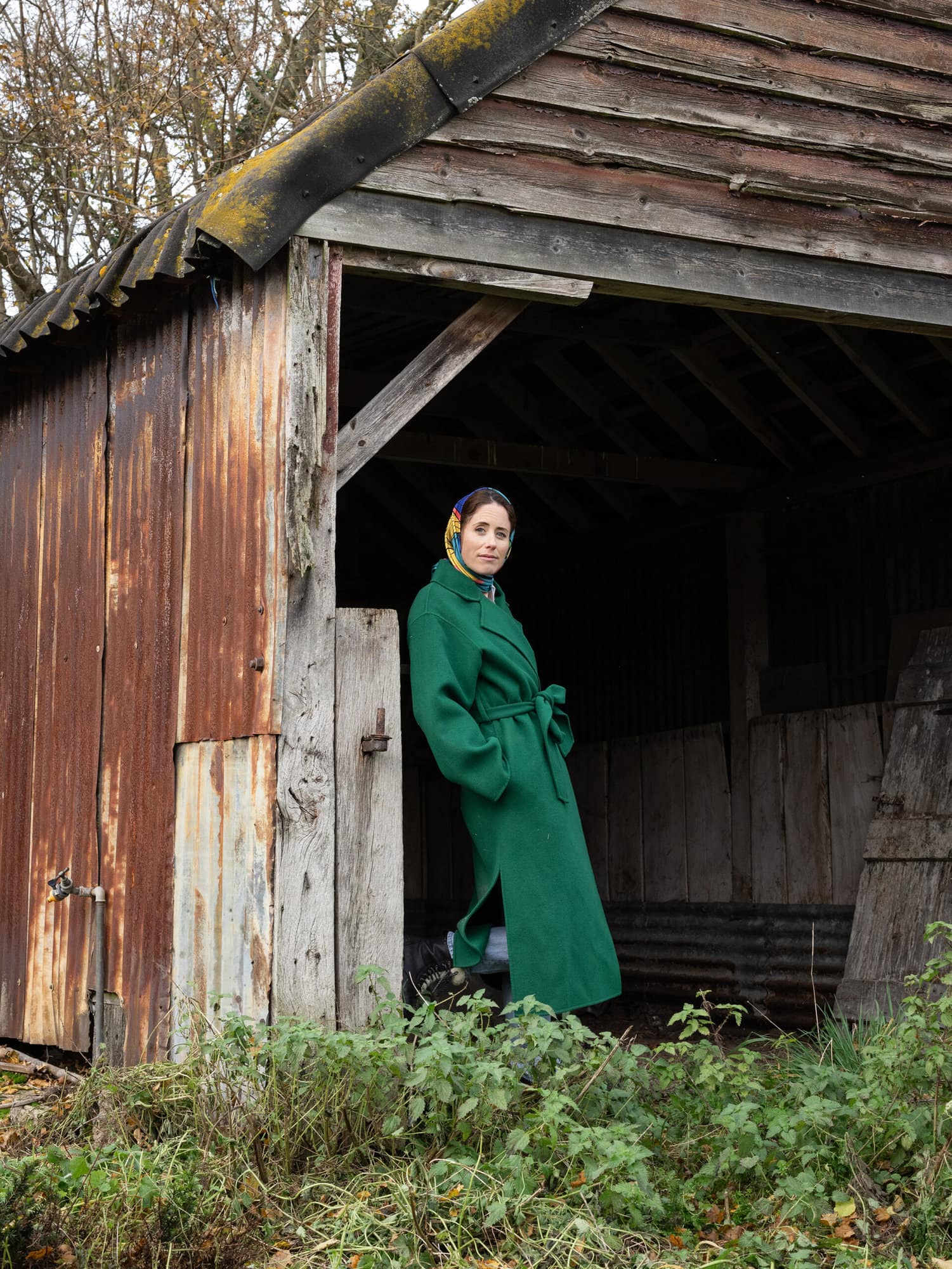 Woman in a green coat and Mai Norman Headscarf leans against a wall of an old barn