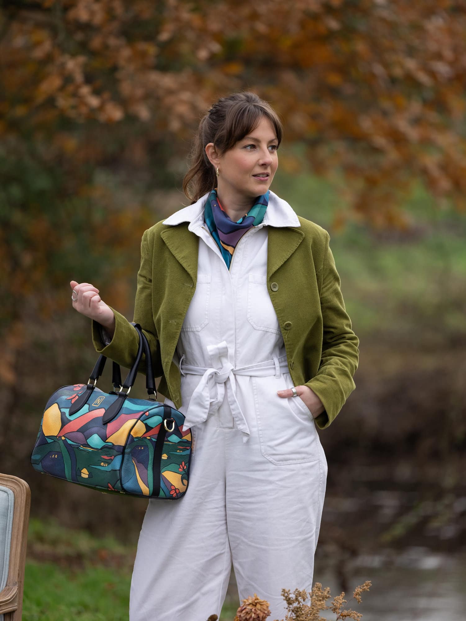A woman in a white jumpsuit stands in the countryside wearing Mai Norman Headscarve and holding a Mai Norman leather bag Photographed by Alison McKenny Photography