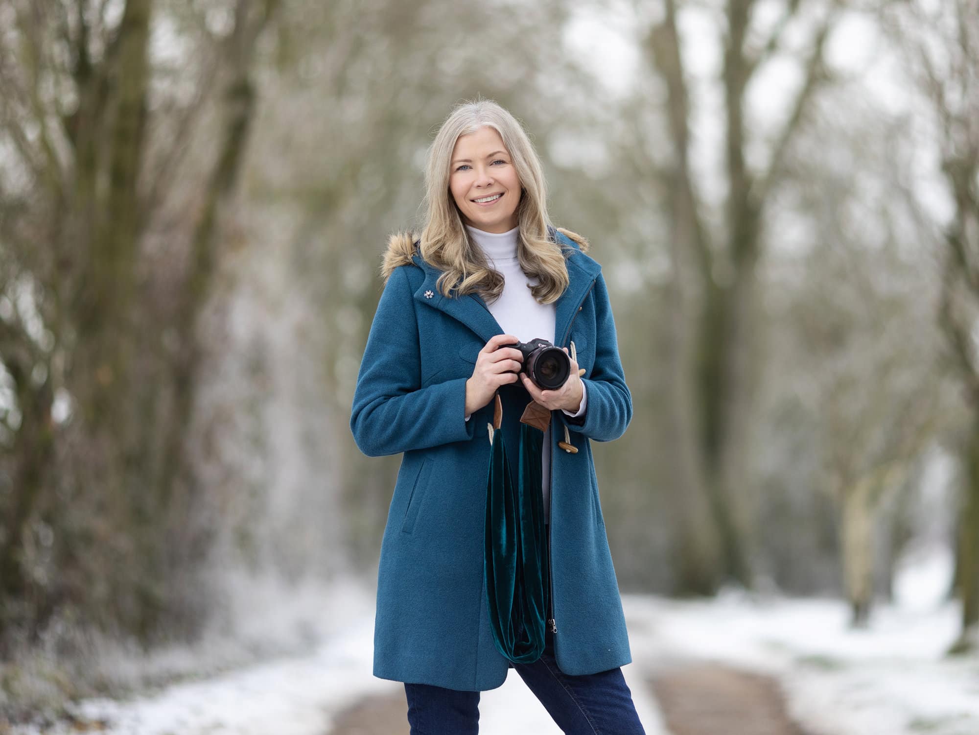 Award winning Portrait PHotographer Alison McKenny stands in the snow with her camera in her hands wearing a teal coat